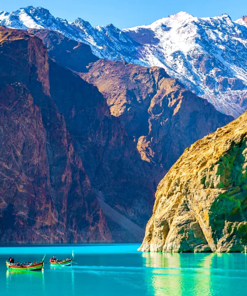 A serene view of Attabad Lake in Gilgit-Baltistan, Pakistan, with a boat gently gliding on its turquoise waters, surrounded by rugged mountains.